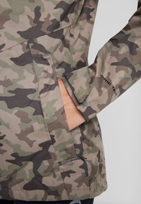 Hand i fickan på en jacka med camouflage-mönster och OMNI-TECH-logotyp på ärmen, närbild av tyg- och söm detaljer.