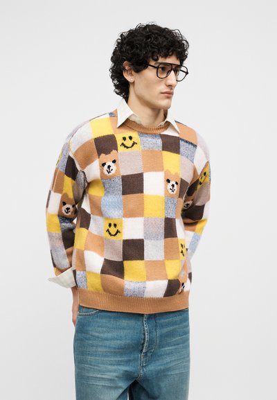 Άνδρας με κατσαρά μαλλιά και γυαλιά που φοράει πολύχρωμο πουλόβερ με καρό μοτίβα, χαμογελαστά προσωπάκια και αρκουδάκια, συνδυασμένο με μπλε τζιν.