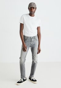 Jean en denim gris avec une coupe droite, associé à un t-shirt blanc uni et des baskets noires. Les accessoires incluent une casquette grise.