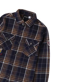 Camicia in flanella di colore blu navy con motivo a quadri marrone e bianco. Presenta due tasche sul petto, chiusura con bottoni e un ricamo sulla manica.