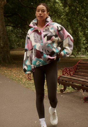 Žena kráčajúca po ceste v parku, oblečená v farebnej vzorovanej flísovej bunde, čiernych legínach a bielych teniskách, v pozadí je drevená lavička a stromy.