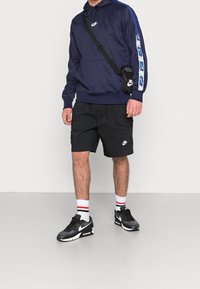 Mies, joka on pukeutunut laivastonsiniseen huppariin, jossa on sinisiä raitoja, mustiin shortseihin, mustiin lenkkareihin, valkoisiin sukkiin ja mustaan Nike-logosin koristeltuun olkalaukkuun.