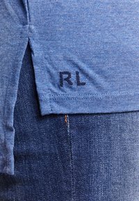 Close-up van de blauwe heathered shirtzoom met zijsplit en geborduurde "RL" initialen bovenop donkere blauw denim jeans.