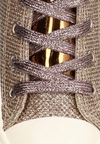 Zapatilla con cordones con un acento metálico dorado, que presenta una parte superior texturizada en champán y cordones texturizados a juego sobre una puntera blanca.