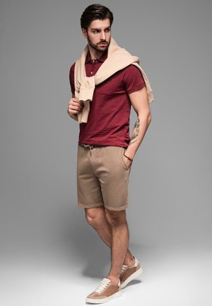 Polo shirt bordeaux, shorts beiges et sneakers beiges. Pull drapé sur les épaules. Comprend des manches courtes et des poches sur les shorts.