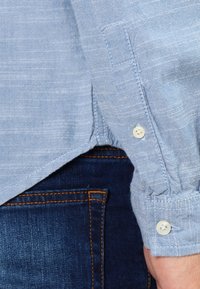 Helles blaues Leinenhemd mit strukturierter Oberfläche, ausgestattet mit Knopfmanschetten und einem abgerundeten Saum. Dunkelblaue Jeans sind teilweise sichtbar.