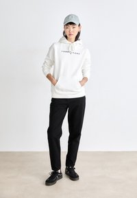 Sudadera blanca con el logo "TOMMY JEANS", bolsillo frontal, combinada con pantalones negros y zapatillas negras, accessorios con una gorra azul claro.