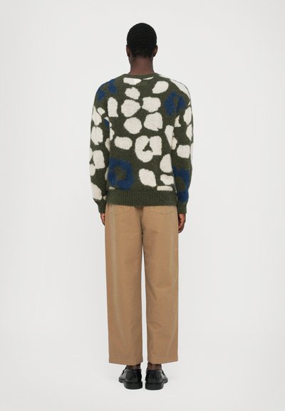 Jersey de punto en verde con patrones abstractos en blanco y azul, combinado con pantalones de pierna ancha beige y zapatos negros. Forma cuadrada, acabado texturizado.