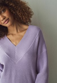 Maglione lilla lavorato a maglia con scollatura a V e texture a coste, caratterizzato da cuciture angolate e maniche ampie su uno sfondo neutro.