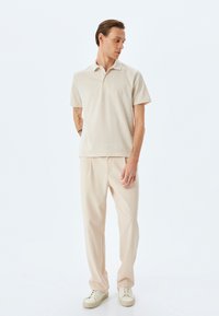 Polo a righe chiare in tono sabbia e crema, abbinato a pantaloni beige larghi e sneakers bianche. Il tessuto appare morbido con una sottile trama.