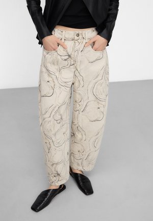 Kobieta ubrana w beżowe spodnie o szerokich nogawkach z czarnym kwiatowym wzorem szkicowym, czarny krótki top, czarną kurtkę oraz czarne buty na kwadratowym nosku.