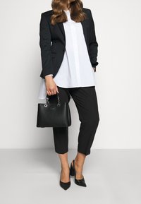 Musta bleiseri valkoisen paidan päällä, muotoon ommellut mustat housut ja mustat teräväkärkiset korkokengät. Pitää kädessään pientä mustaa käsilaukkua, jossa on ketjuviimeistelyä.