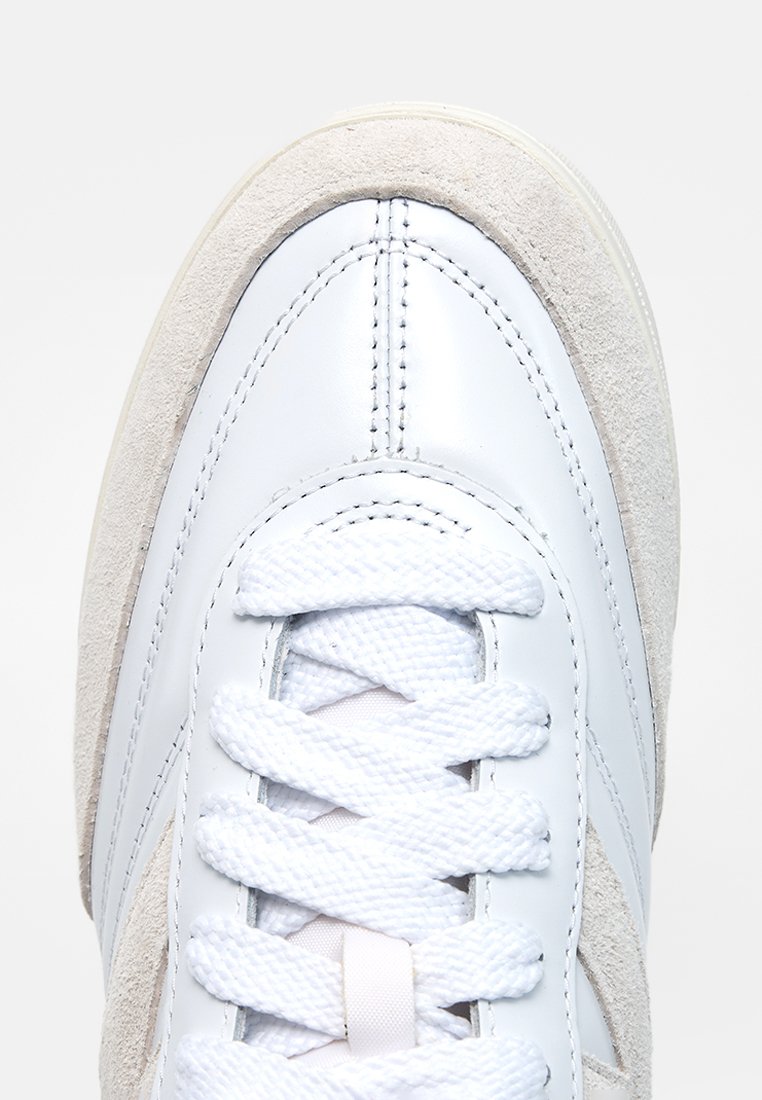 Punta de zapatilla blanca con cordones blancos texturizados y ribete de ante beige, costuras detalladas visibles en la parte superior de cuero liso.