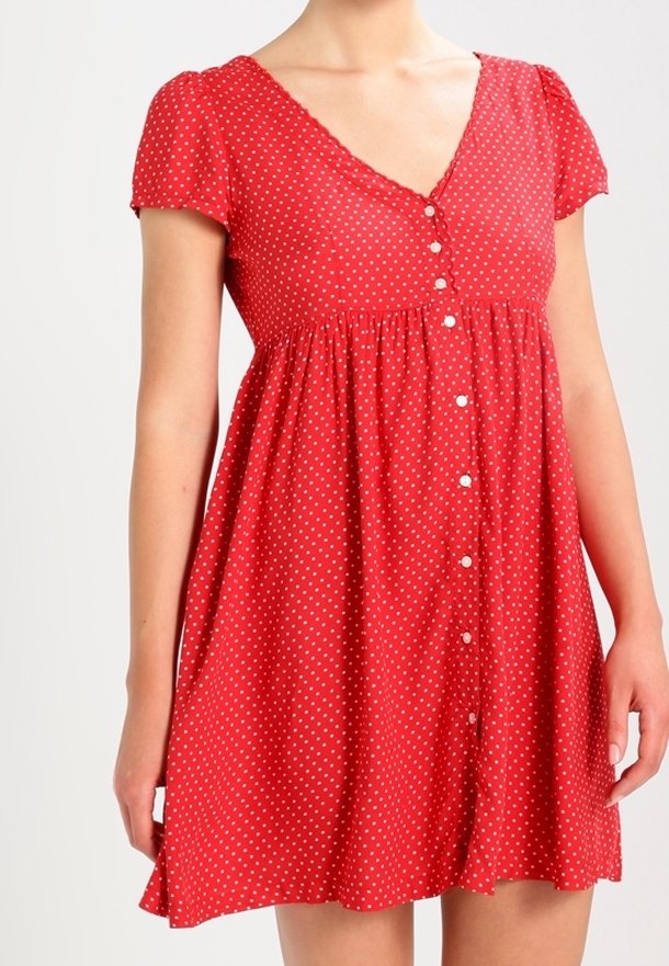 Röd kortärmad klänning med V-ringning, prydd med vita prickar och knäppning fram. Utrullande design, lättviktsmaterial och samlad midja.