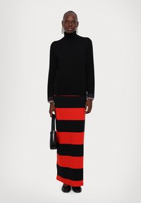 Femme portant un col roulé noir et une longue jupe rayée rouge et noire, tenant un sac à main noir, se tenant devant un fond uni.