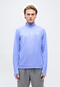 Άνδρας με ανοιχτό γαλάζιο πουλόβερ με φερμουάρ έως τη μέση και γκρι παντελόνι, στέκεται μπροστά σε απλό λευκό φόντο.