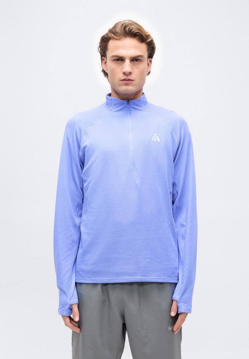 Άνδρας με ανοιχτό γαλάζιο πουλόβερ με φερμουάρ έως τη μέση και γκρι παντελόνι, στέκεται μπροστά σε απλό λευκό φόντο.