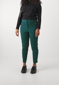 Green tailored trousers with a slim fit, featuring side pockets and a textured finish, paired with a black long-sleeve top and black shoes.