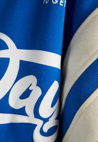 Niebieska bluza z białym napisem, z dużym logo w skrypcie i kremowym akcentem. Materiał wydaje się miękki z teksturowanym wykończeniem.