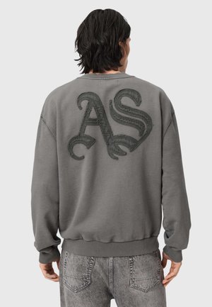 Personne aux cheveux foncés portant un sweat-shirt gris avec de grandes lettres texturées « AS » au dos et un jean gris clair.