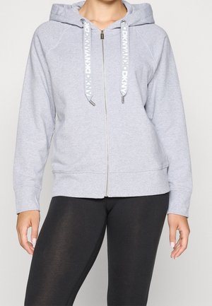 Vrouw draagt een lichtgrijze hoodie met rits met het DKNY-logo op de koordjes en zwarte legging, staand met ontspannen handen langs het lichaam.