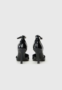 Crne lakirane cipele s visokom potpeticom, s kaišem oko gležnja, šiljastim nosom i sjajnom završnom obradom. Imaju usku, stiletto potpeticu.