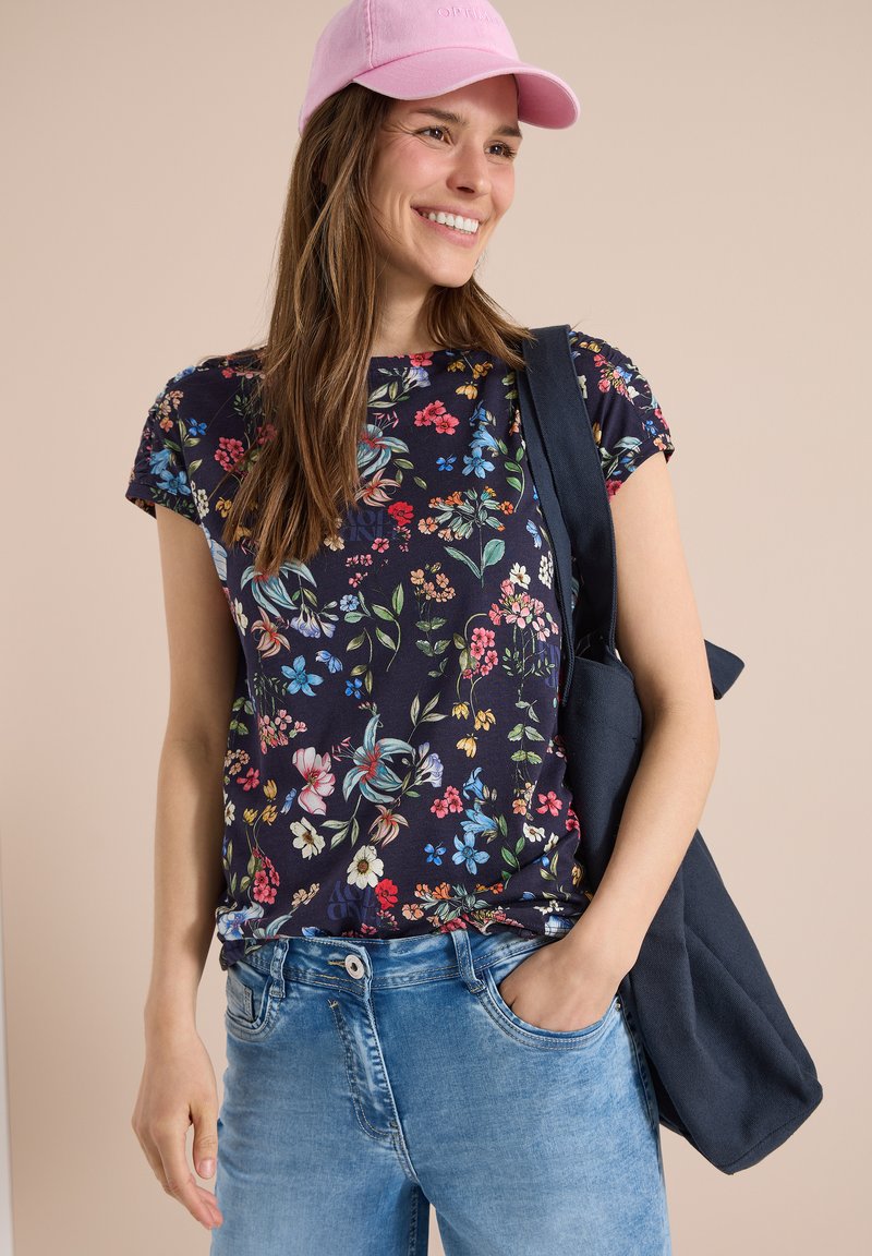 Mujer sonriente con gorra rosa, camisa azul marino con flores, vaqueros azul claro y un bolso azul marino colgado del hombro sobre un fondo beige.