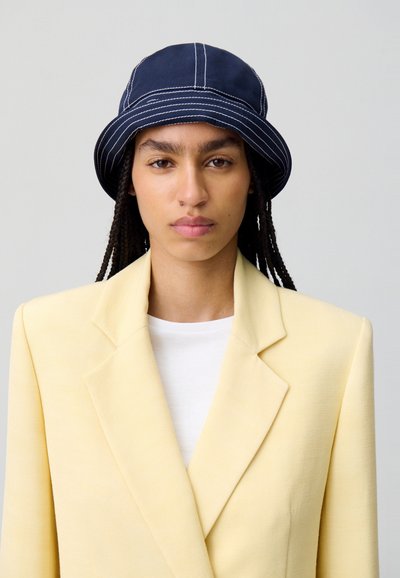 Cappello a secchiello blu navy con cuciture bianche, abbinato a una giacca leggera gialla e una camicia bianca. Texture liscia, design strutturato.
