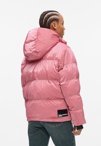 Pink puffer jacket with horizontal quilted design and a hood. Made of shiny material, features black logo patch on the lower left side.