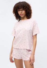 Kobieta z kręconymi włosami w jasnoróżowym krótkim topie w kwiaty i pasujących krótkich spodenkach, stojąca na tle jednolitego białego tła.