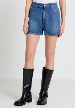 Donna che indossa pantaloncini di jeans blu e stivali neri in pelle fino al ginocchio, in piedi contro uno sfondo chiaro e semplice.