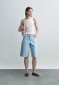 Žena stojaca v svetloružovom tielku bez rukávov, svetlomodrých rifľových šortkách po kolená, plochých topánkach s leopardím vzorom a sivej kabelke na rameno s strapcami.