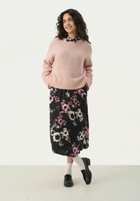 Un pull en rose clair, flou, avec un ourlet côtelé, associé à une jupe midi noire à motif floral avec des accents rose et crème, et des chaussures noires.