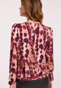 Kobieta o jasnobrązowych włosach nosi bluzkę z długim rękawem w różowe, fioletowe i musztardowe abstrakcyjne kwiatowe wzory, połączoną z czarnymi skórzanymi spodniami.