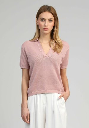 Jeune femme aux longs cheveux blonds portant un haut en maille rose à manches courtes et un pantalon plissé blanc, debout avec une main dans la poche.