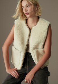 Creme faux-shearling vest med lynlås foran, rund krave og to forlommer, lagd over en hvid top og parret med mørke jeans.