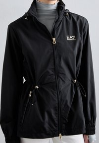 Svart zip-up jacka med EA7 Emporio Armani-logotyp, justerbara midjesnören, bärs över ljusgrå polotröja och vita byxor.