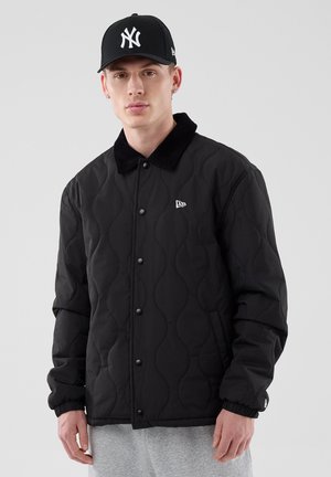 Joven con una chaqueta acolchada negra con botones a presión y una gorra de béisbol negra de los New York Yankees, de pie frente a un fondo claro y liso.