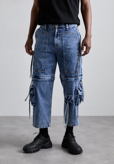 Denim cargo pants with multiple pockets, drawstring details, mid-blue wash, and a relaxed fit. Paired with black sneakers and ankle socks.