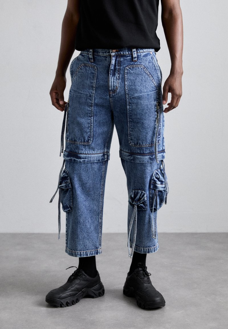 Pantaloni cargo din denim cu multiple buzunare, detalii cu șnur, spălare de culoare albastru mediu și croială lejeră. Asortați cu adidași negri și șosete până la gleznă.