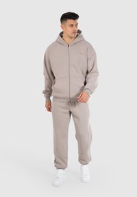 Beige Kapuzenpullover mit Reißverschluss und passende Jogginghose, aus weichem Stoff, mit seitlichen Taschen und gerippten Bündchen, kombiniert mit weißen Sneakers.