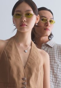 Dos modelos llevan gafas de sol con tintes amarillos. El modelo de frente viste una blusa sin mangas marrón con botones, mientras que el otro lleva una camisa a cuadros.
