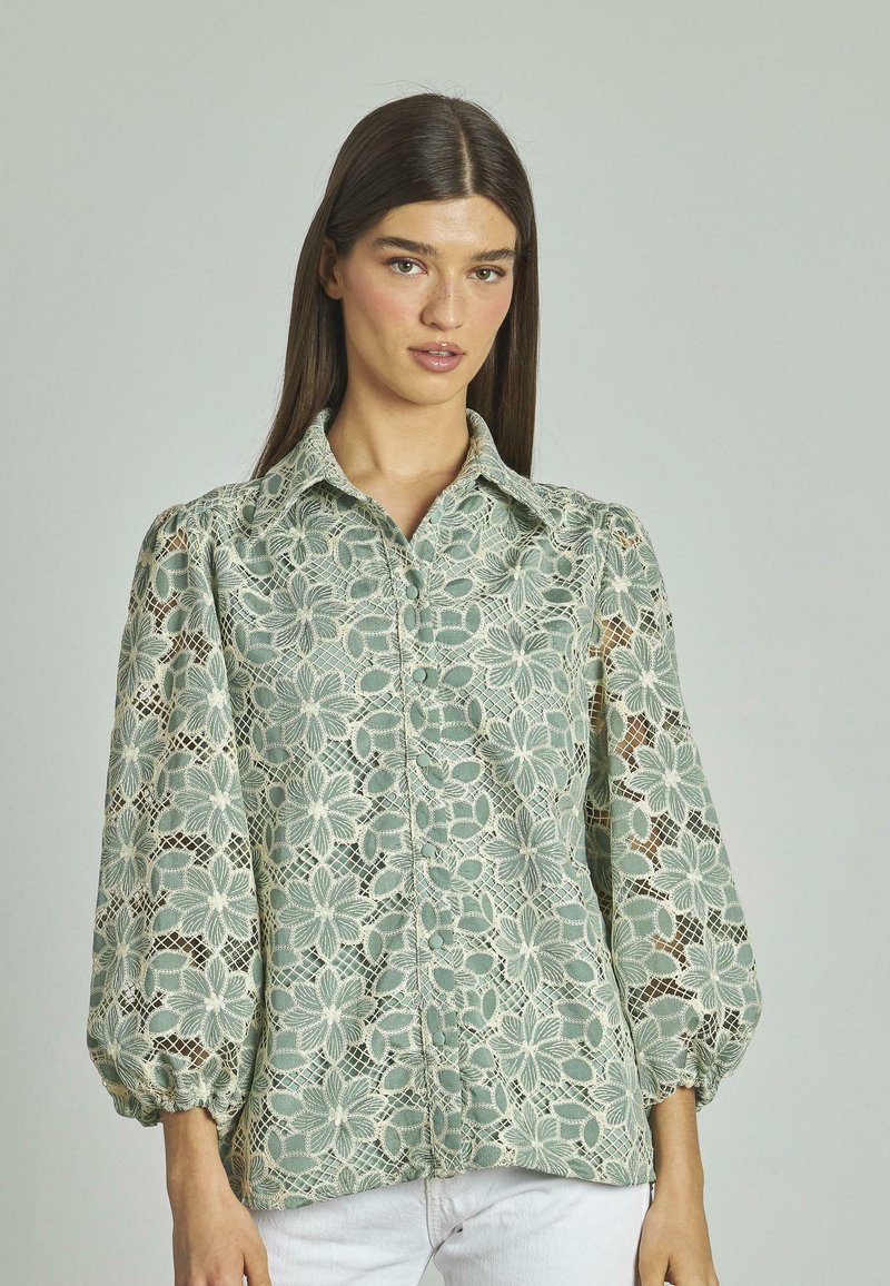 Blusa in pizzo di un verde morbido con motivi floreali, chiusura con bottoni anteriori e maniche a palloncino con polsini elasticizzati. Tessuto arricchito con dettagli intricati.
