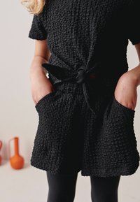 Sort struktureret jumpsuit med korte ærmer, en knyttet talje og sidelommer, lavet af indsamlet stof. Bæret med sorte leggings.