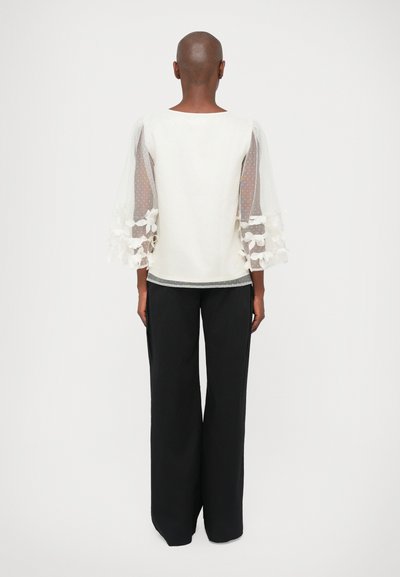 Bela majica s prosojnimi teksturiranimi rukavima koji imaju cvjetne aplikacije, u kombinaciji s crnim širokim hlačama. Jednostavni okrugli izrez i detalji na hemu.
