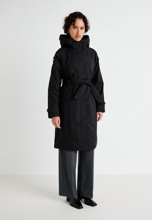 Abrigo largo negro con cintura ajustada, con capucha, cremallera frontal, bolsillos laterales y tela lisa e impermeable. Combinado con pantalones de pierna ancha.