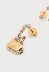 Χρυσά σκουλαρίκια σε σχήμα καρδιάς με μια λαμπερή σειρά από κρυστάλλους και χαραγμένο λογότυπο "GUESS", με κυρτό σχεδιασμό και καρφίτσα πίσω μέρος.