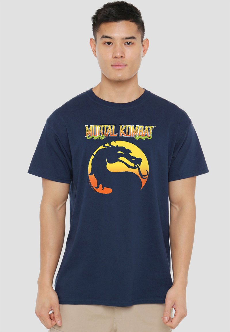 Young man wearing a navy blue T-shirt with a yellow and orange Mortal Kombat dragon logo and text on the chest, standing against a plain background.