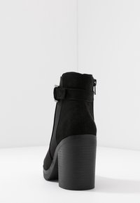 Botín de ante negro con tacón alto, que presenta una cremallera lateral y una correa ajustable. Textura suave con una suela de goma resistente.
