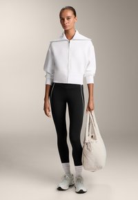 Felpa bianca a zip corta con colletto alto, abbinata a legging neri con piping laterale. Il modello tiene una borsa beige chiara.